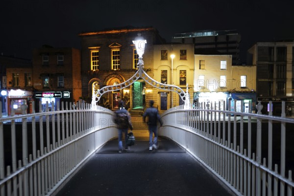 A view of two adults crossing the Ha'penny Bridge on the River Liffey near Temple Bar at night-time. Dublin, Ireland, Europe
