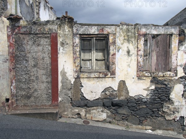 An old ruined house partly collapsed on a sloping street with a blocked up door crumbling walls and fading red painted windows with closed shutters