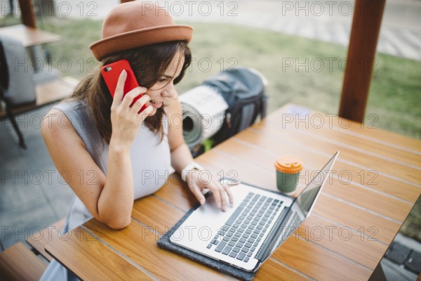 Theme tourism and travel young student. beautiful young Caucasian girl in dress and hat sits in street cafe at wooden table using laptop technology and mobile phone. Search and book hotel and ticket