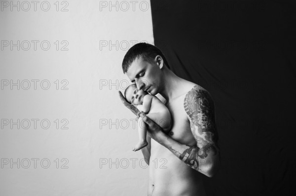 Father with tattoo on his arm is holding his newborn baby on a black and white background.