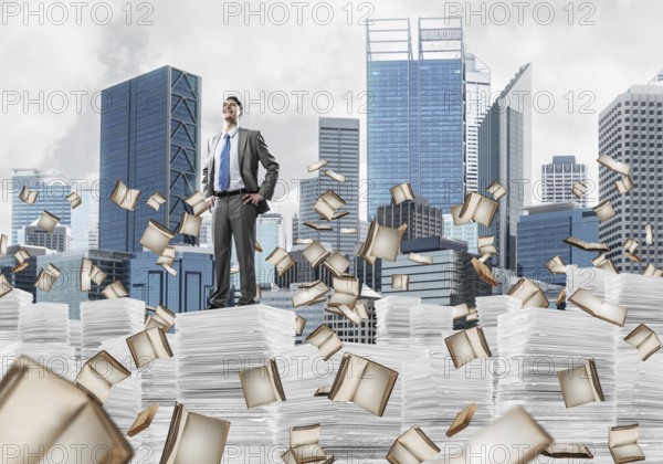 Confident businessman in suit standing on pile of documents among flying books with cityscape on background. Mixed media