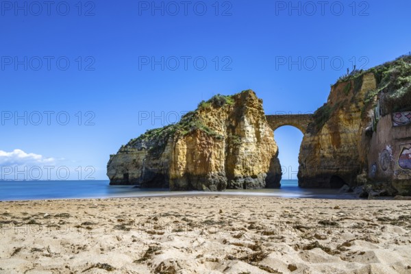 Yellow rocks and cliffs on a sandy beach. A bay by the sea, over which the old stone bridge of the Pinhão fortress connects two cliffs. Praia da Caldeira, Lagos. Algarve, Portugal, Europe