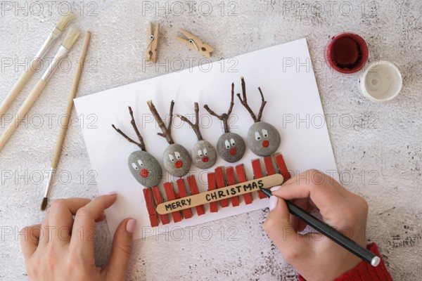 Diy Merry Christmas deer on greeting card postcard made of pebbles, sea stones, clothespins and branches on white background. Natural Eco style diy. Gift idea. Step by step. Top view. Process kid children craft. Do it yourself