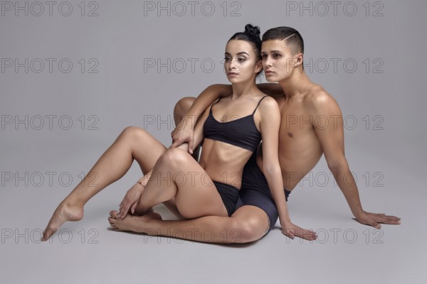 A pair of a graceful athletic ballet dancers in black suits are posing over a gray studio background. Good-looking male in black shorts is embracing a lovely woman in a black swimwear. Ballet and contemporary choreography concept. Art photo