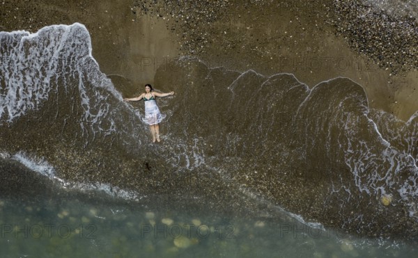 Woman with swimsuit and white dress resting on a sandy beach with breaking waves on the shore. Overhead shot. Aerial drone photograph
