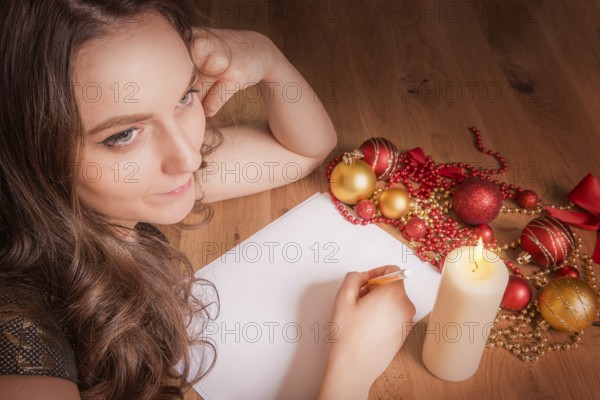 Beautiful smiling woman portrait, thinking about what to write with pencil on blank paper, at candlelight and surrounded by Christmas decorations