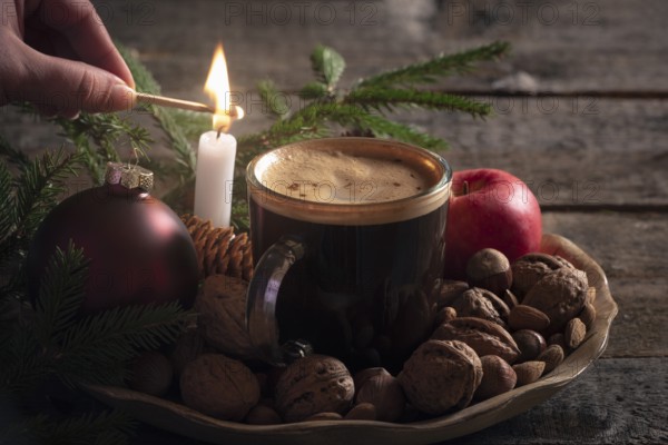 Christmas cozy atmosphere with a woman lighting a candle on a wooden platter full with nuts, apples and a cup of coffee, in low light