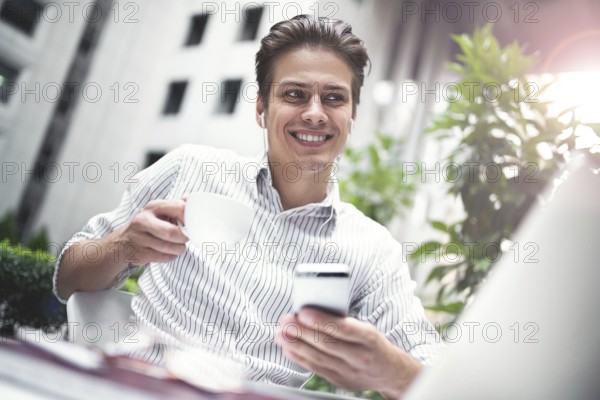 Happy handsome businessman looking at computer screen while typing something on laptop keyboard during work break outdoors, businessman with headphones looking at the notebook sitting in cafe working