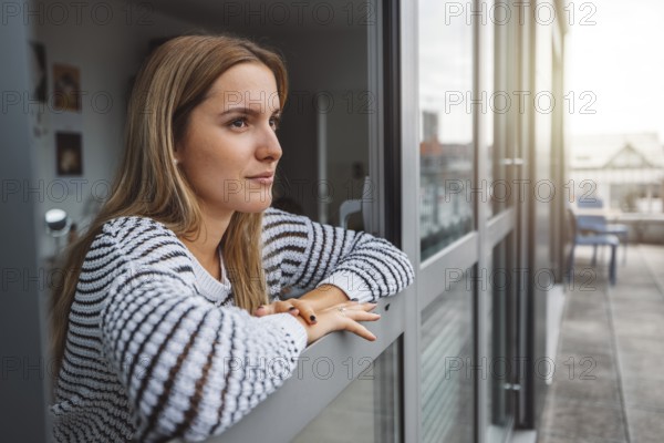 Young blonde caucasian woman looking out the window of her dorm room getting some fresh air on a cold autumn day
