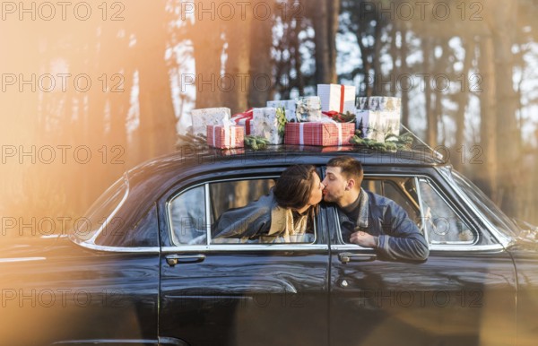 Loving man and woman sticking out of retro car windows and kissing with Christmas gifts on roof in park