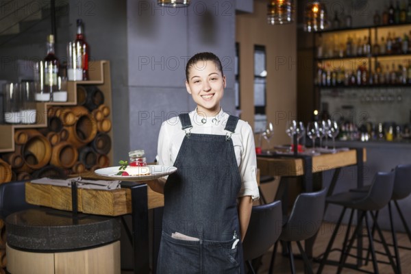 Ganson, officers, beautiful deushka with a smile and gathered hair in working form apron accepts an order in a restaurant standing near the servered wooden tables