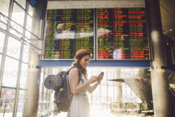 Theme travel and tranosport. Beautiful young caucasian woman in dress and backpack standing inside train station or terminal looking at a schedule holding a red phone, uses communication technology