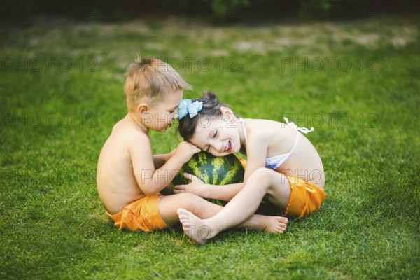 Two children, Caucasian brother and sister, sitting on green grass in backyard of house and hugging big tasty sweet watermelon berry, green striped colour. Theme healthy food and ecological product
