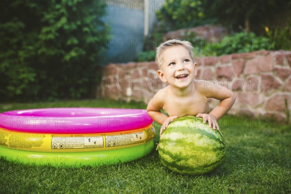 Toddler cute Caucasian white European race boy sitting hugging holding huge watermelon and smiling on background of green graß, backyard of the house and a round children pool on a sunny summer day