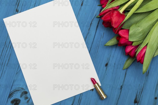 Red tulips, lipstick and a white sheet of paper for your greetings on the background of blue wooden board