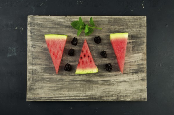WATERMELON BLACKBERRY AND MINT ON THE BACKGROUND. THREE PIECES OF RIPE JUICY WATERMELON WITH RIPE BERRY BLACKBERRY MINT LIE ON A WOODEN BOARD ON A DARK BACKGROUND