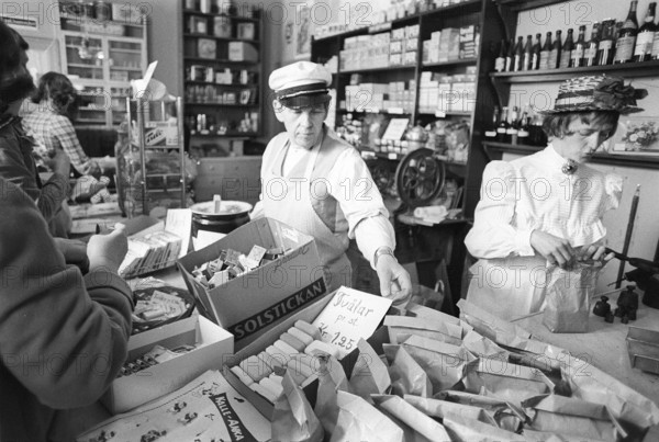 A general store museum in the 1970s. Photograph taken inside a general store, capturing its appearance from 100 years ago, are displayed for the public who shop there. The images show a nostalgic scene where customers purchase goods over the counter, served by a shopkeeper standing behind it, selling wares in a traditional, hands-on manner. The setup evokes a bygone era of commerce, blending history with the present as modern shoppers experience the same space once depicted in the century-old snapshots. Kristoffersson ref 8-22
