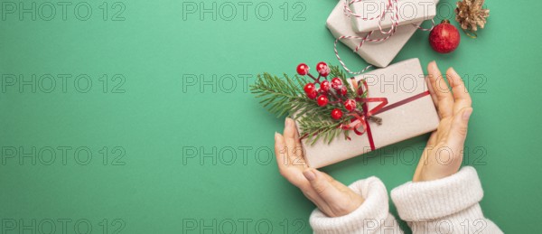Christmas or New Year celebration green paper festive background with female hands holding wrapped present box, decoration fir tree, cones, berries, sparkly red balls. Space for text, food photography, food photography