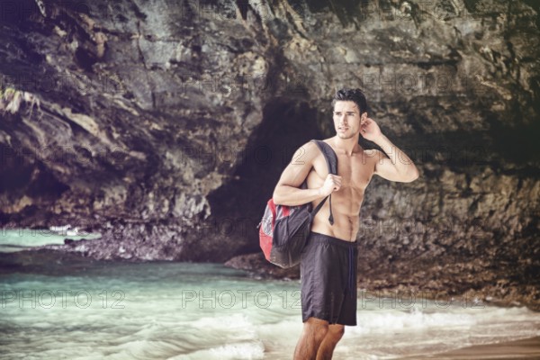 Half body shot of a handsome young man standing on a beach in Phuket Island, Thailand, shirtless wearing boxer shorts, showing muscular fit body, with backpack on one shoulder, Asia