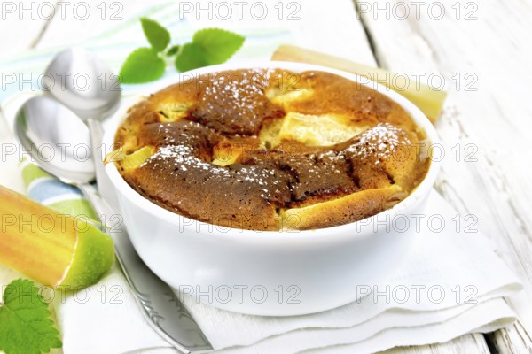 Pie clafouti with rhubarb in the bowl, mint and rhubarb stems on a napkin on wooden plank background