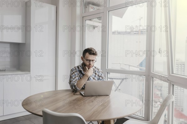 Man in a checkered shirt thoughtfully working on a laptop in a bright workspace