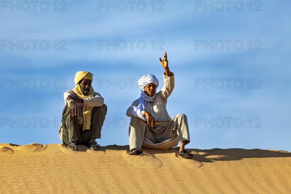 Two men sit on a sand hill in the desert under a clear blue sky, two Tuareg sit on a sand dune and talk in the Sahara in Algeria