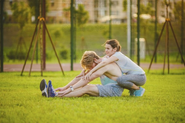 Young in love heterosexual Caucasian couple, man with beard woman with big breasts doing exercise stretching muscles, warming up outdoor training in park on graß Girl coach is teacher and mentor