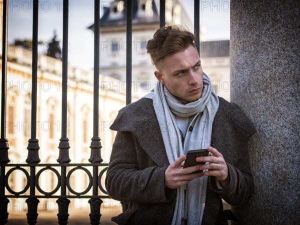 Handsome trendy man wearing dark coat standing and looking down at a cell phone that he is holding, outdoor in European city setting with elegant old historic building behind