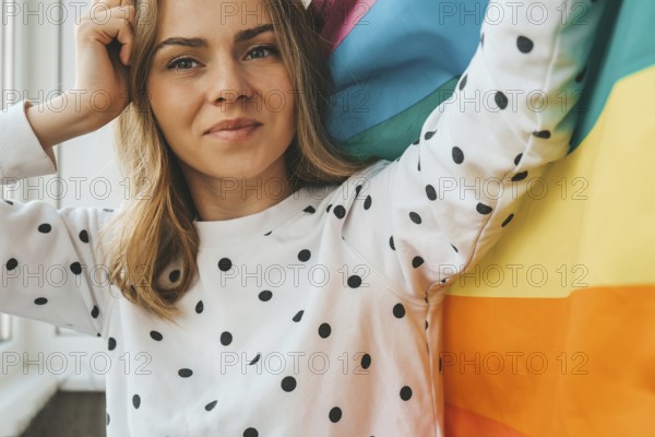 Young blonde millennial hippie woman with rainbow LGBTQ flag at home balcony. Peace and freedom the symbol of LGBT, spending and sharing loving time. Support LGBTQ community. Young lesbian woman