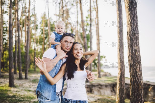 Family mom, dad and daughter sits at the daddy on the shoulders, and parents kiß on the nature in the forest in the summer at sunset