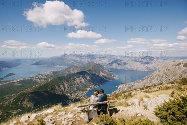 Bride and groom are sitting on a bench and looking at the Kotor bay. Excellent view from the mountains of Montenegro.