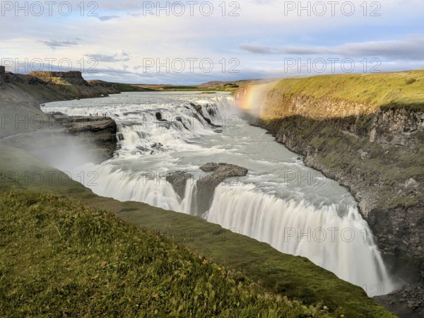 Majestic waterfall cascading over cliffs with rainbow and lush greenery under a cloudy sky. Iceland