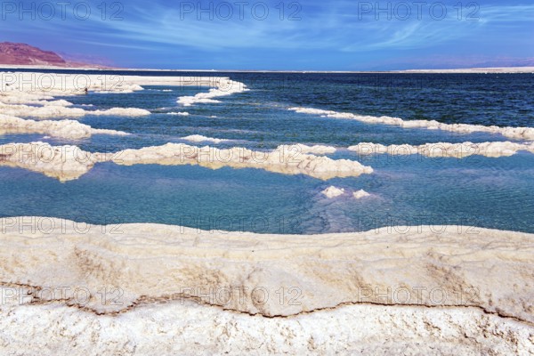 The evaporated salt forms bizarre patterns on the surface of the water. Cirrus clouds fly in the blue sky. Windy spring day. The Israeli coast of the Dead Sea