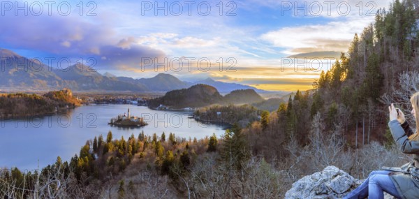 Panorama with the lake Bled under a colorful winter sky at sunrise and a young woman enjoys the view sitting on a rock