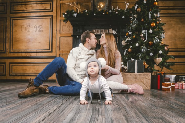 Theme family holiday New Year and Christmas. Young caucasian family mom dad son 1 year sit wooden floor near fireplace christmas tree on Christmas evening. Baby learning walk creeps background parents