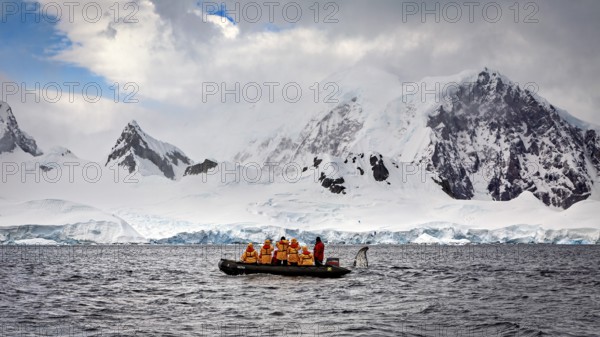 Group in a boat in the foreground, surrounded by snow-covered mountains and overcast sky, people in a rubber dinghy travelling on the sea in the Southern Ocean in Antarctica