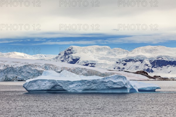 A large iceberg floats in the sea, surrounded by snowy mountains under a blue sky, iceberg in the Southern Ocean in Antarctica