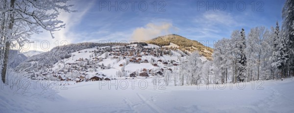 Panoramic winter scene of snow covered La Clusaz village nestled in a hilly landscape in Annecy, France. Wooden cabins are scattered across the snowy hillside, surrounded by snow laden trees