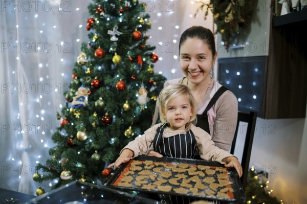 Smiling mother with little daughter sitting at the table with a baking sheet of raw Christmas cookies.