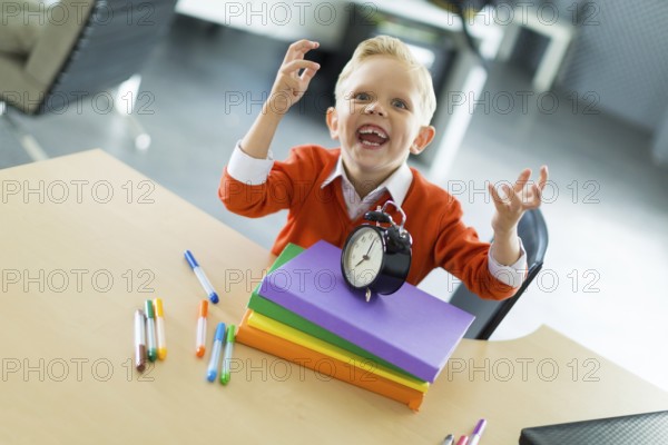 Cute blonde caucasian kid sit at the table with colorful folders, hold alarm clock, evil laugh, look at camera Blurred office background, focus on lad