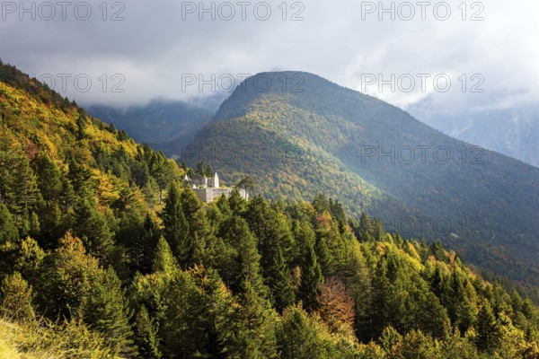 Travel to Slovenia. Ruins of a medieval castle on a mountainside. The mountain road passes through yellow and orange trees and grasses. The Julian Alps