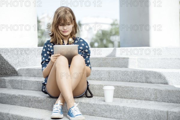 Beautiful young woman with vintage music headphones around her neck and a cup of coffee to go, surfing the internet on tablet PC and sitting on the stairs against urban city background