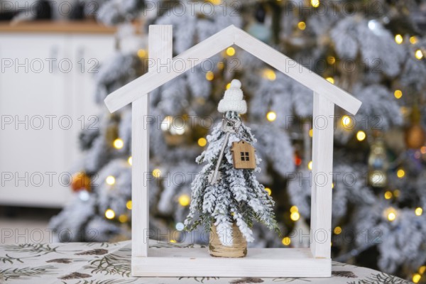 Key on Christmas tree and tiny house on cozy home with Christmas decor on table of festive white kitchen. Gift for New Year. Insurance, moving to new house, mortgage, rent and real estate