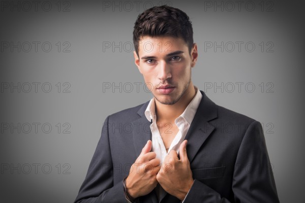 Young businessman confidently posing in front of camera, wearing business suit without neck-tie, with shirt open on neck, on dark background