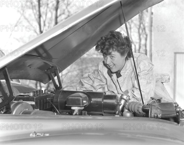 In the 1950s. A woman in an overall fixing something on the car that has it's hood open. Sweden 1950. Kristoffersson ref 40K-8