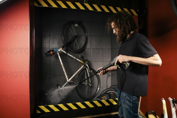 Services professional washing of a bicycle in the workshop. Close-up of hand Young Caucasian stylish man doing bicycle cleaning using automatic electric water pump. Sprays scatter from the pressure