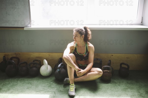The girl is resting, taking a break during training in the gym. Sits by the window near a lot of weights