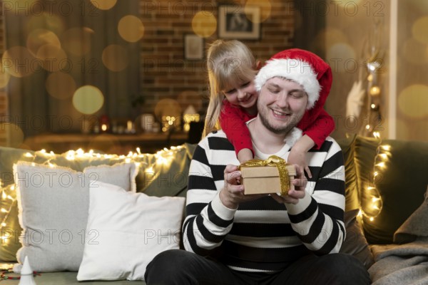 Merry Christmas and Happy New Year. Christmas gift surprise. A little smiling daughter girl hugging father back holding gift indoors at home decorated for holiday. Handsome man is holding giftbox