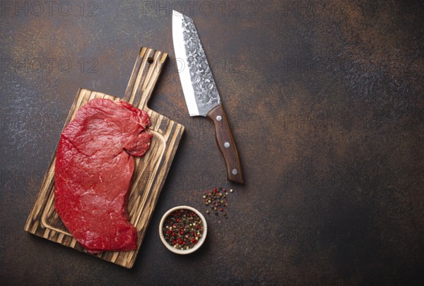 Raw uncooked top round beef steak on wooden cutting board with big kitchen knife and pepper on dark brown rustic stone background top view, cooking meat steak with copy space, food photography, food photography