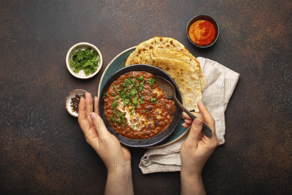 Female hands holding a bowl and eating traditional Indian Punjabi dish Dal makhani with lentils and beans served with naan flat bread, fresh cilantro on brown concrete rustic table top view, food photography, food photography